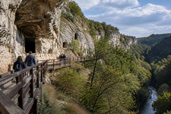Visitors on a timber walkway beneath pale limestone cliffs above a wooded canyon, early in the day, Rock-Hewn Churches of Ivanovo