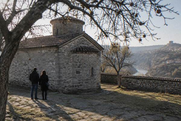 A couple stopping in front of a low thick-walled stone church in a walled yard under fruit trees, Church of the Nativity