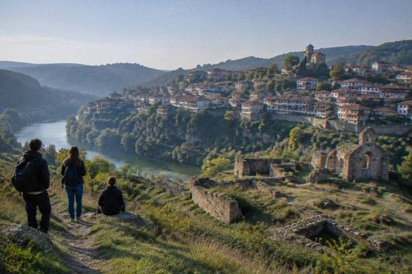 A small group on a grassy hillside path looking across a river gorge at the stepped houses of an old town, early in the day, Trapezitsa