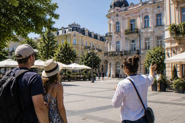A guide and two visitors on a broad pedestrian square of ornate old buildings, Ruse