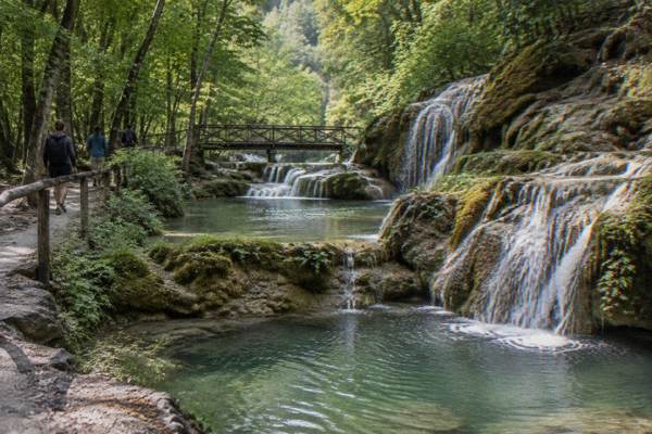 Walkers on a forest path beside pools of green water, Krushuna Falls