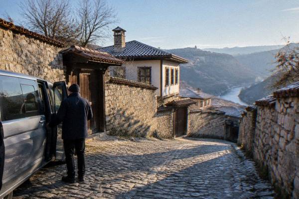 A driver-guide opening the door of a minivan on a quiet village lane lined with tall stone walls, Arbanasi