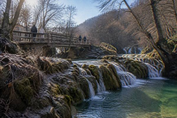 Visitors on a wooden footbridge above clear water cascading over mossy terraces, Krushuna Falls