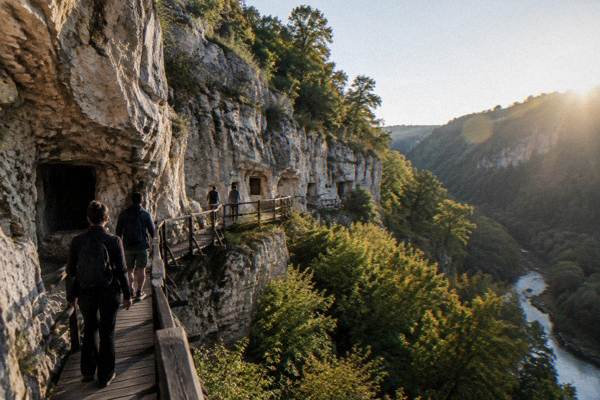 Visitors on a timber walkway beneath pale limestone cliffs above a wooded canyon, Rock-Hewn Churches of Ivanovo