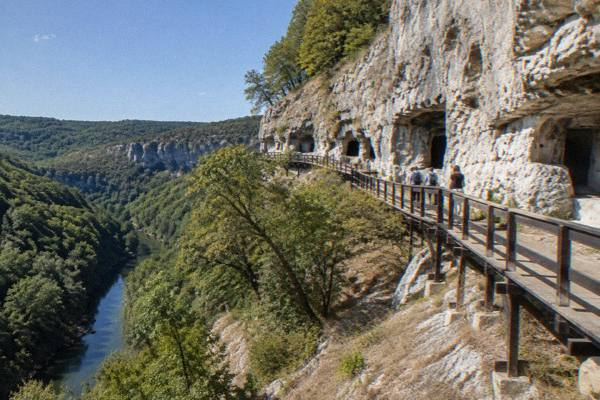 Visitors on a timber walkway beneath pale limestone cliffs above a wooded canyon, Rock-Hewn Churches of Ivanovo