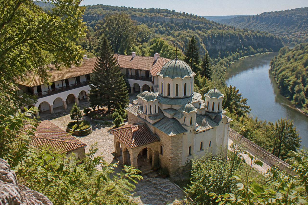 A Bulgarian Orthodox monastery church with white walls and green domes seen from a wooded hillside above a river gorge