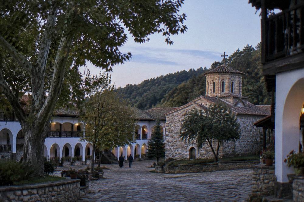 A monastery courtyard with a stone path, plane trees and arched monastic wings under a calm sky