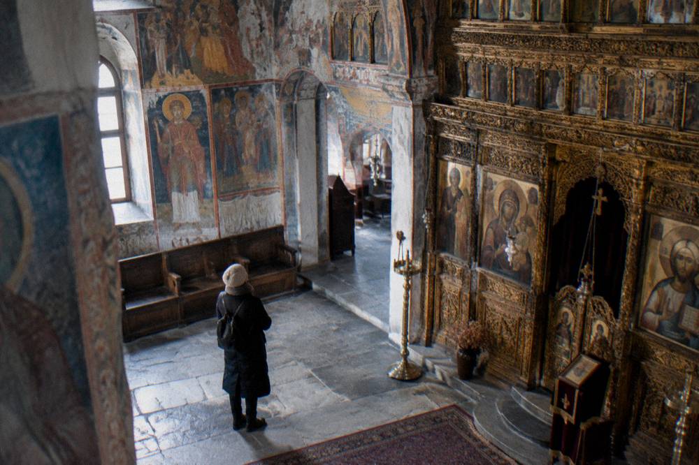 A visitor standing quietly in a monastery church nave looking up at painted walls and a carved iconostasis