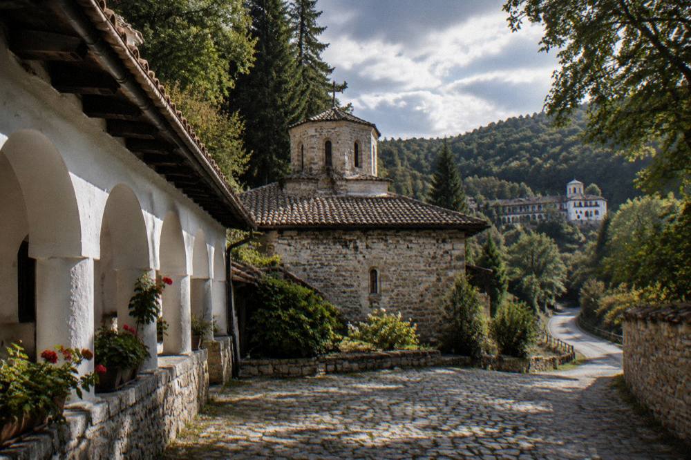 A quiet country road winding through wooded hills towards a monastery complex in the distance