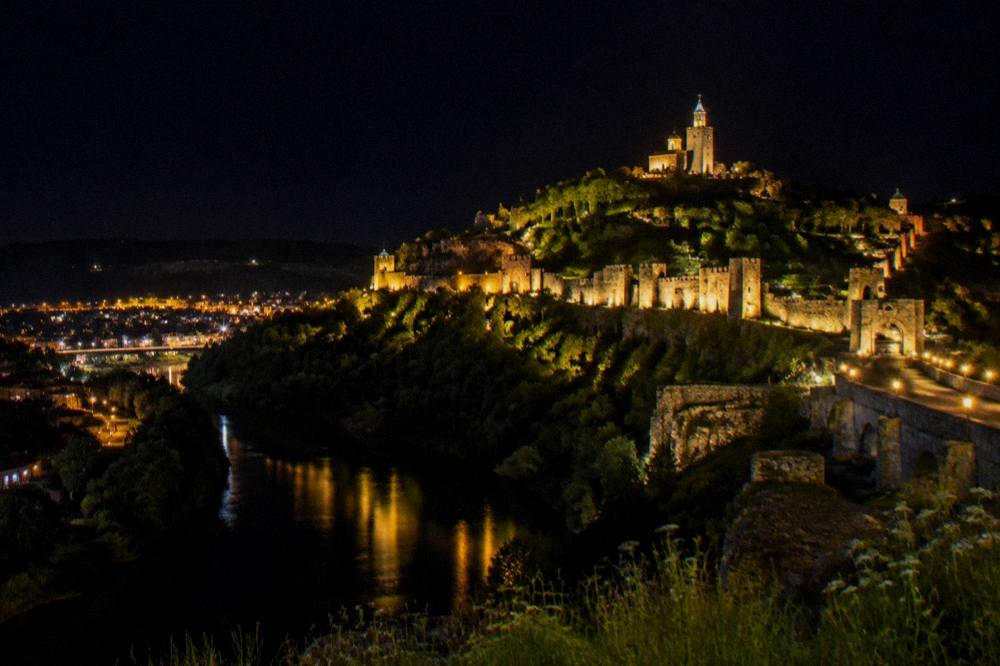 A night panorama of a hilltop fortress glowing above a dark river bend and city lights