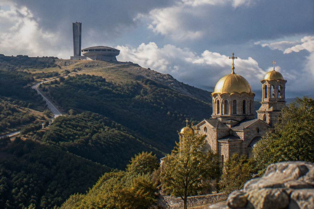 A stone church with golden domes standing below forested mountain slopes