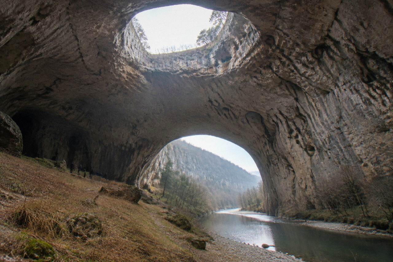 The wide open arched entrance of a large cave above a river, with daylight streaming in