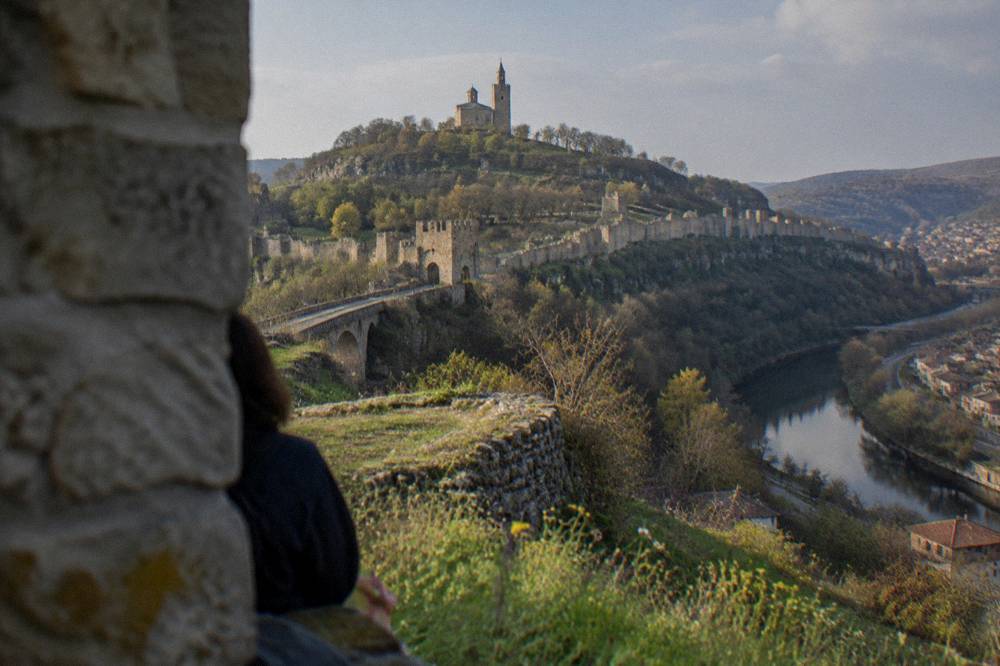 A stone fortress wall on a hill above a winding river town at dusk