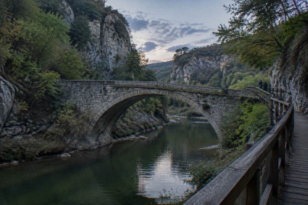 A stone arch bridge over a small river with cliffs and trees in the background under a partly cloudy sky
