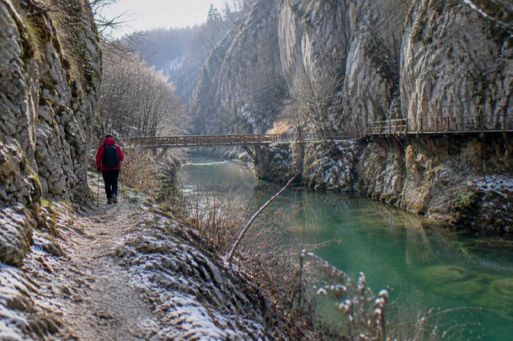 A hiker in a red jacket walking a dirt trail beside a turquoise river between grey cliffs