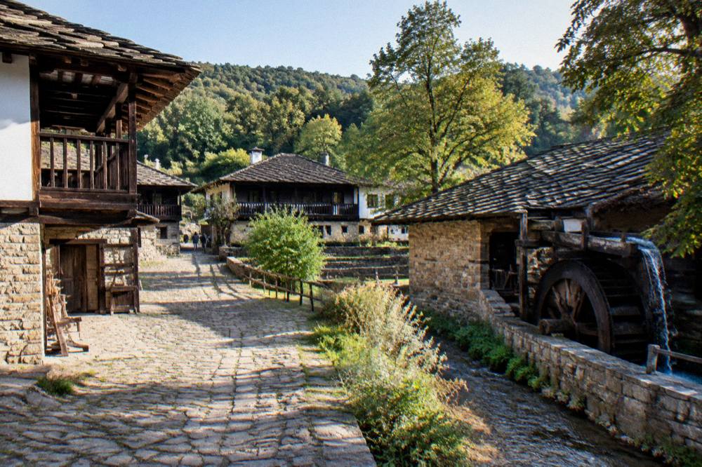 Wide view of a museum courtyard with craft houses, trees and hills in late afternoon light
