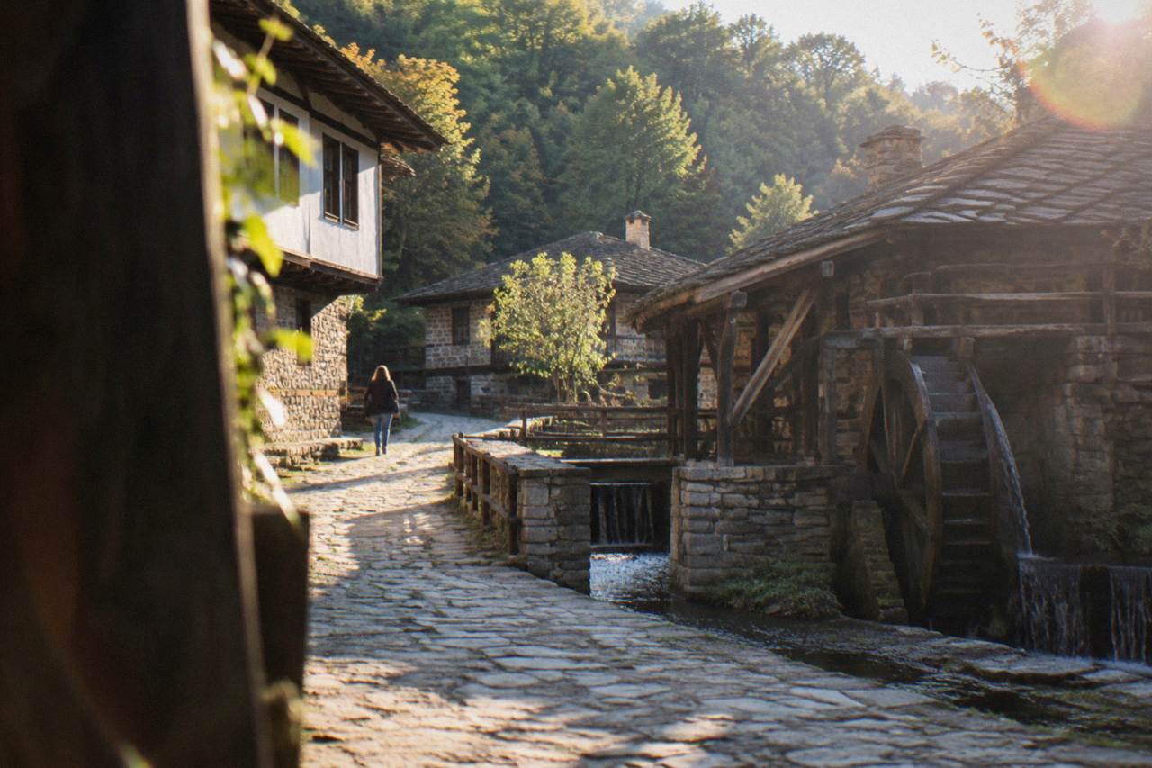 Cobbled street of a Bulgarian revival-era open-air museum with old stone houses and a water wheel