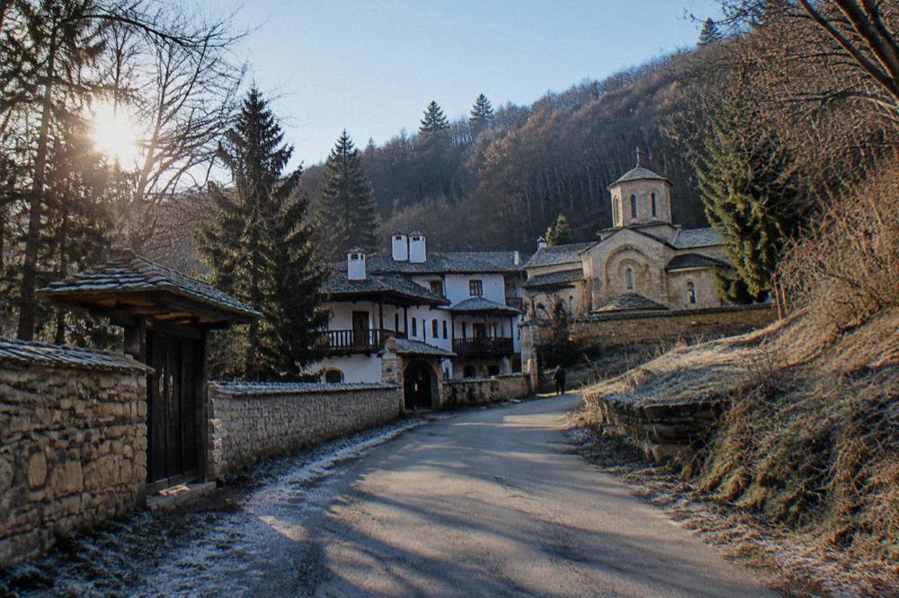 A quiet village road running between wooded slopes toward a monastery building