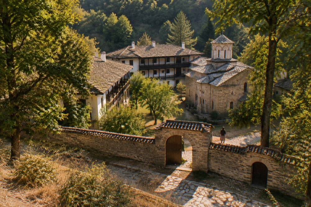 A stone monastery wall with arched gates and trees casting shade
