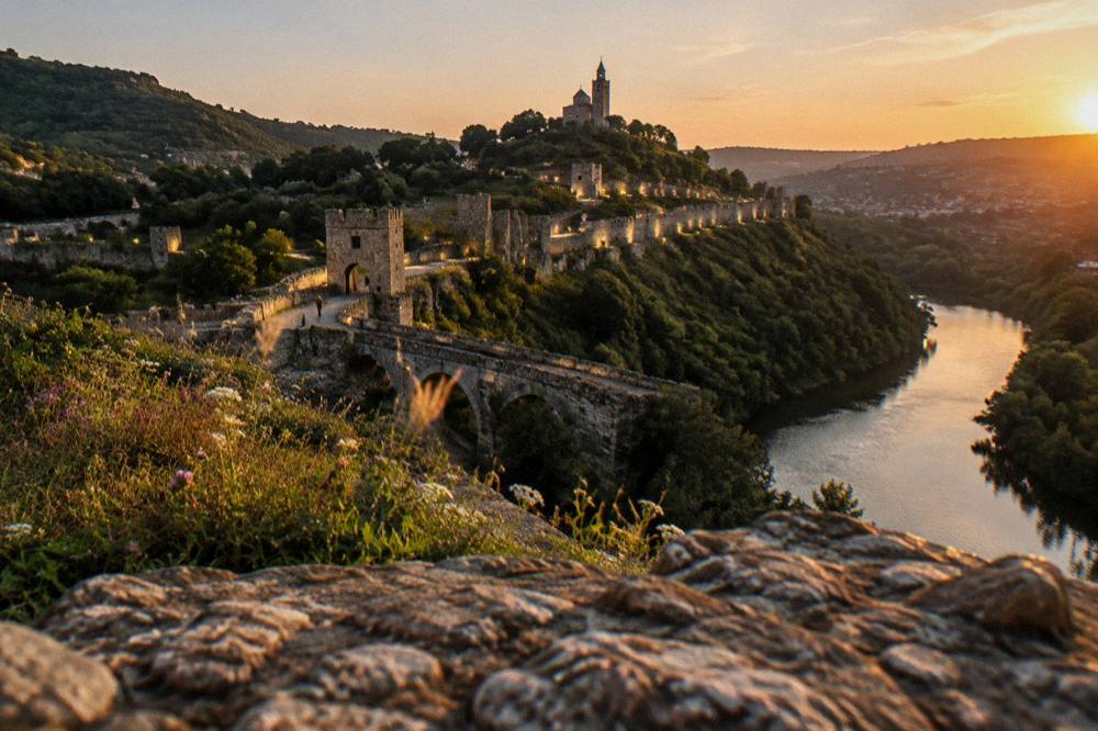 A stone fortress wall on a hill above a river bend at dusk with lit paths