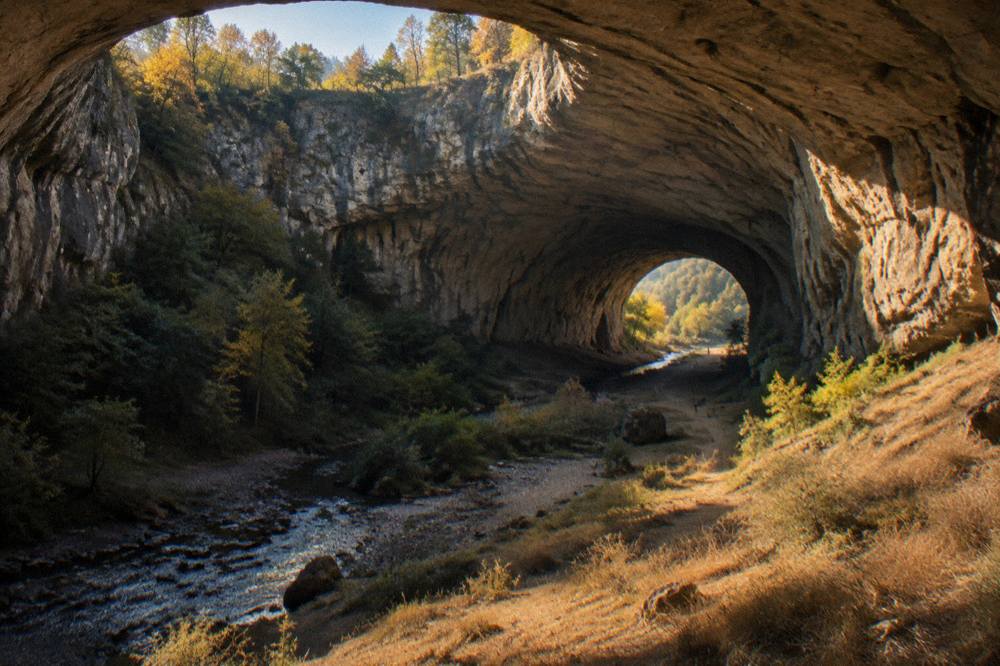 A wide cave passage lit by natural daylight with a person-sized silhouette at the entrance