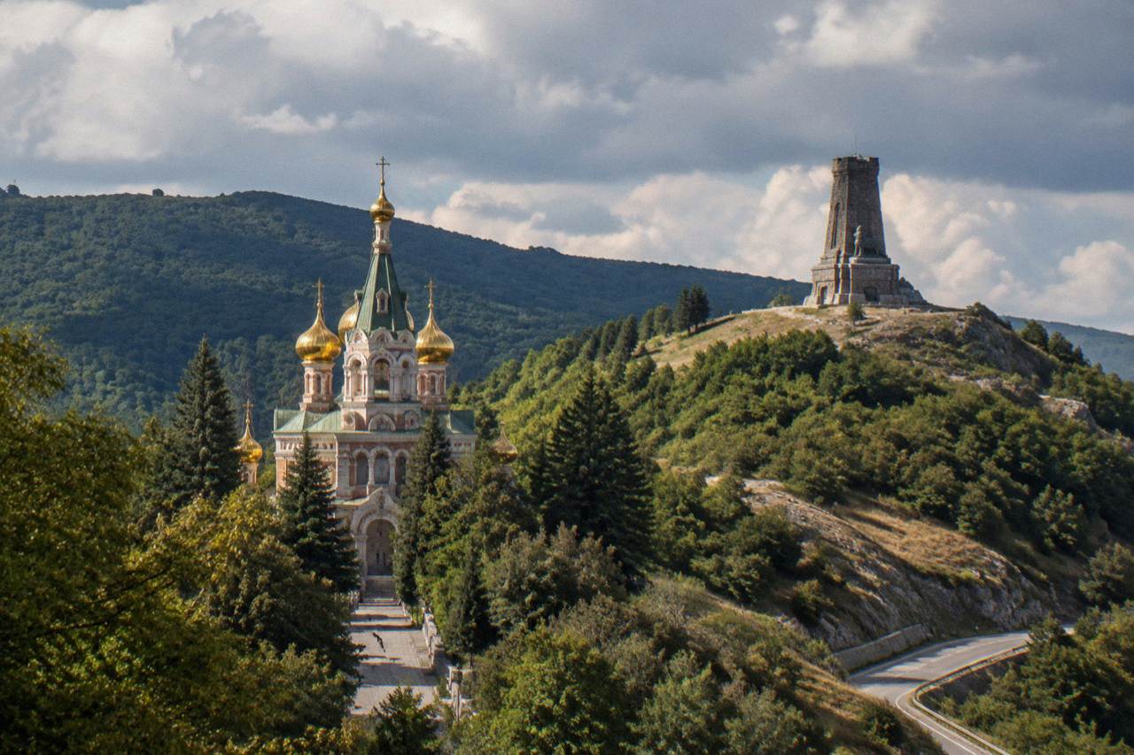 Stone memorial tower standing on a green mountain ridge above a winding mountain road