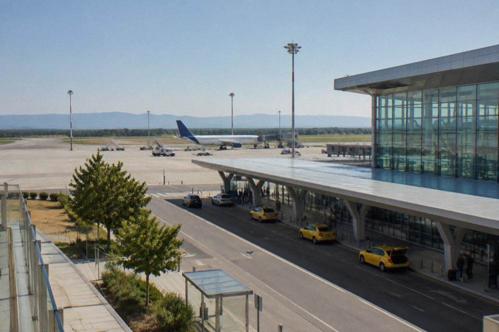 An airport terminal exterior under a clear sky with a parked airliner in the background