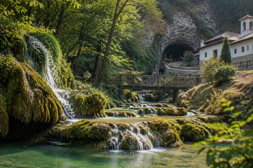 A cave entrance in a wooded hillside beside monastery buildings