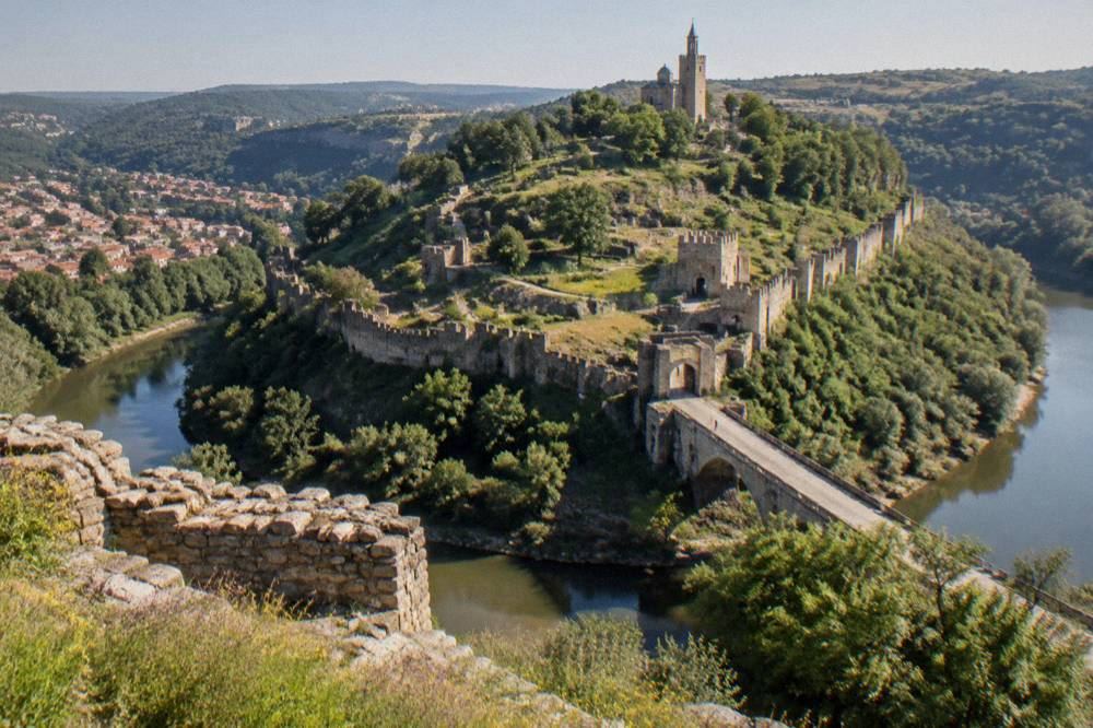 A stone fortress wall on a hilltop above a river bend seen from a viewpoint