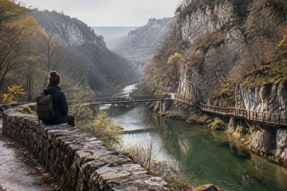 A hiker with a small backpack resting on a stone wall above a river valley