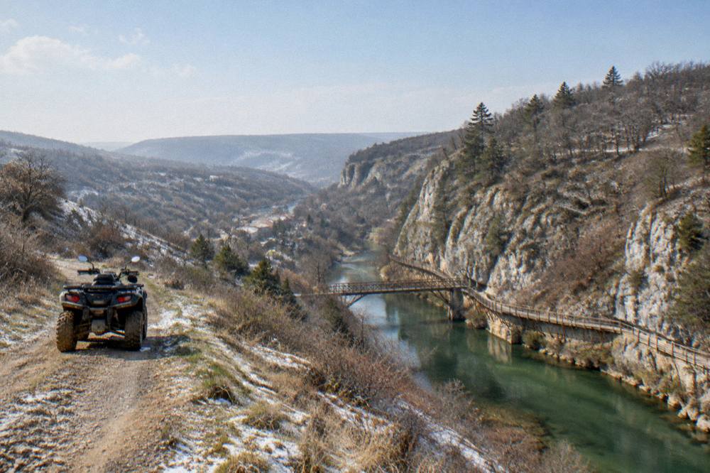 An all-terrain vehicle on a dirt track through open hills above a river town