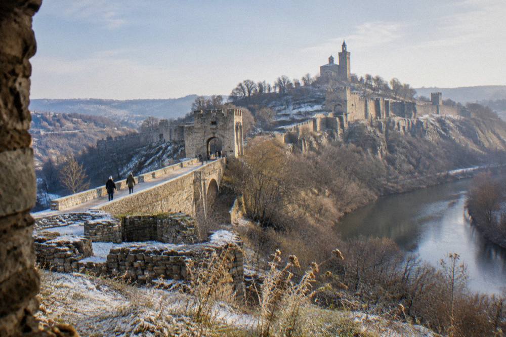 A stone fortress wall and tower rising above a river bend under a pale winter sky